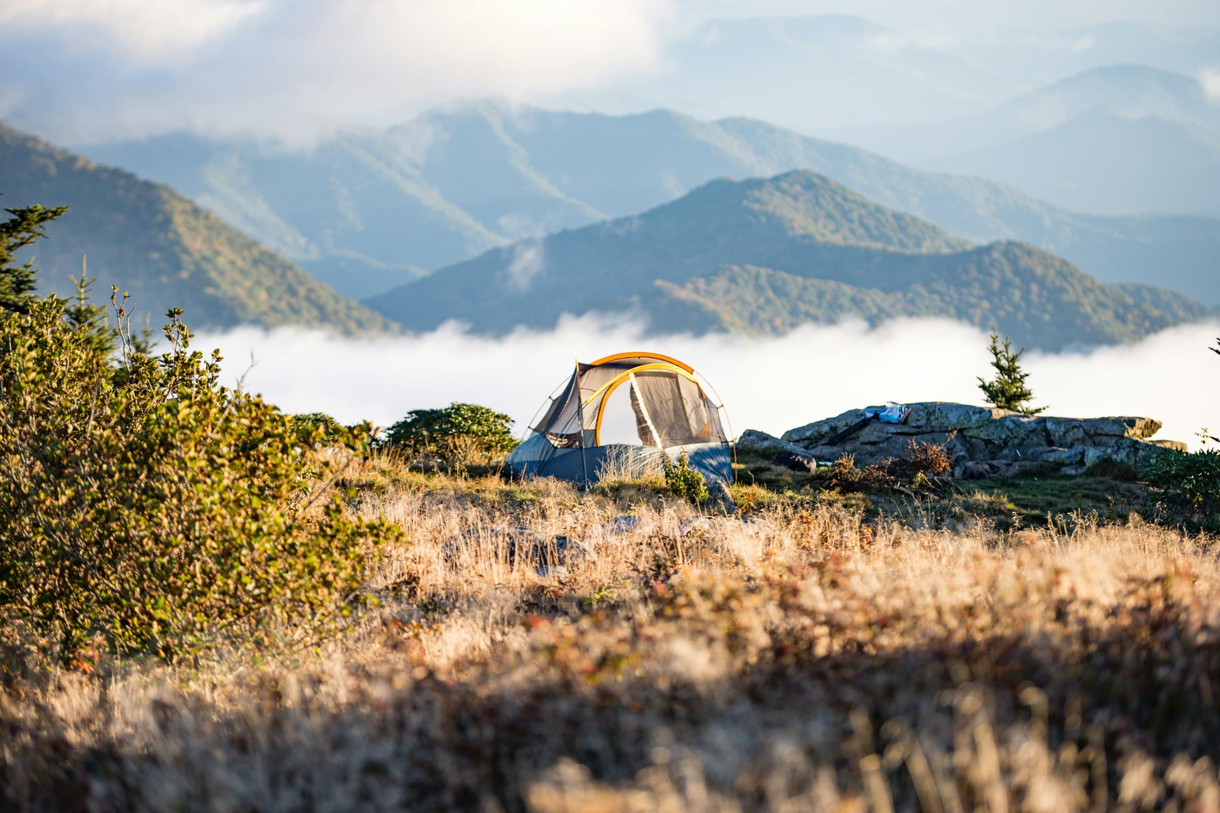 A dome tent on a grassy ridge above a bank of low cloud, with layered mountains fading into haze behind it.
