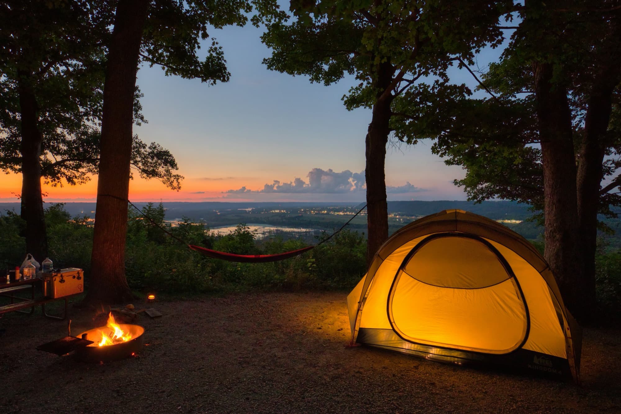 A lit dome tent and a campfire at a hilltop campsite at dusk, with a river valley and orange sky beyond.
